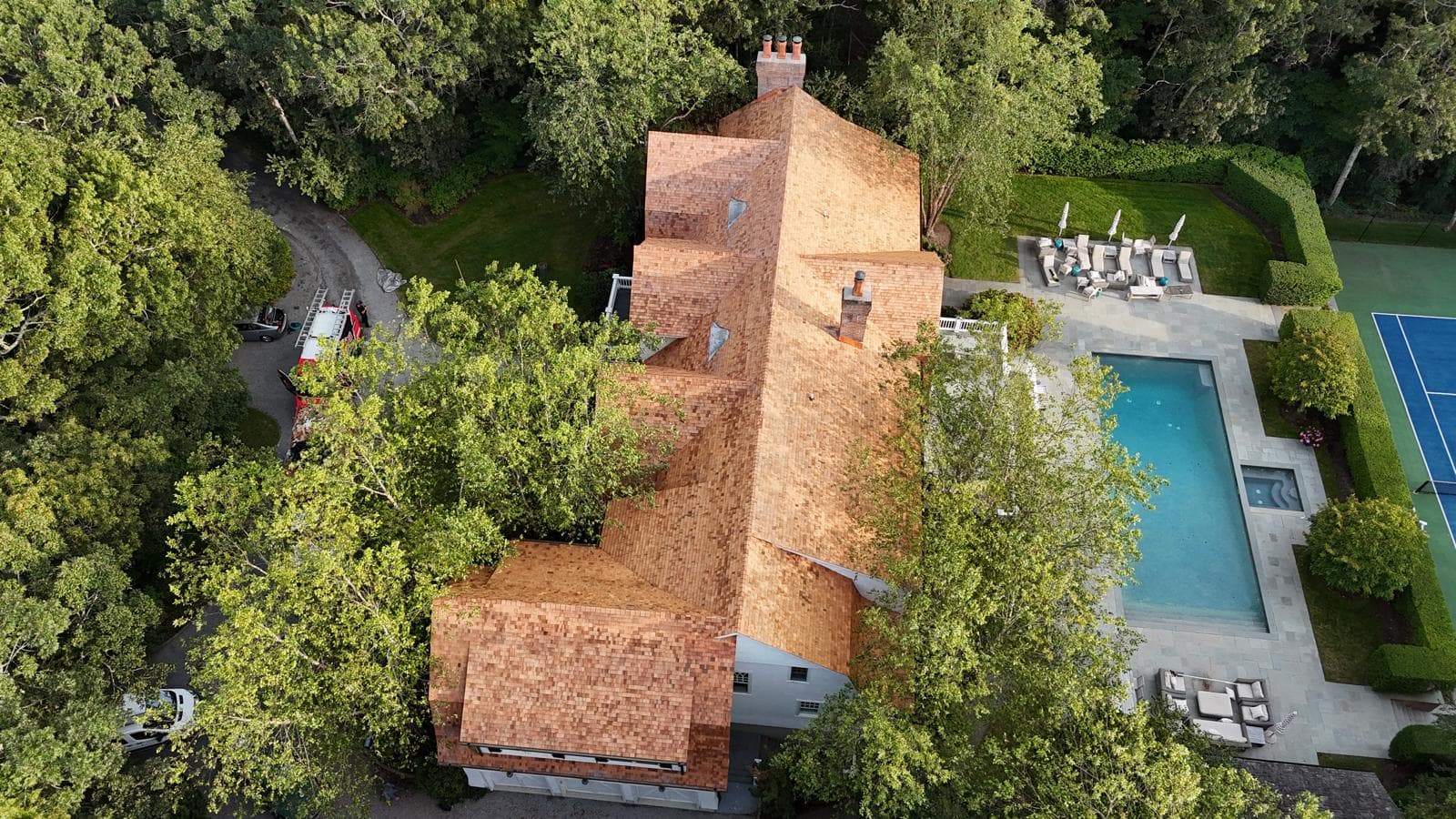 Aerial view of a long cedar shake roof beside a pool and a tennis court
