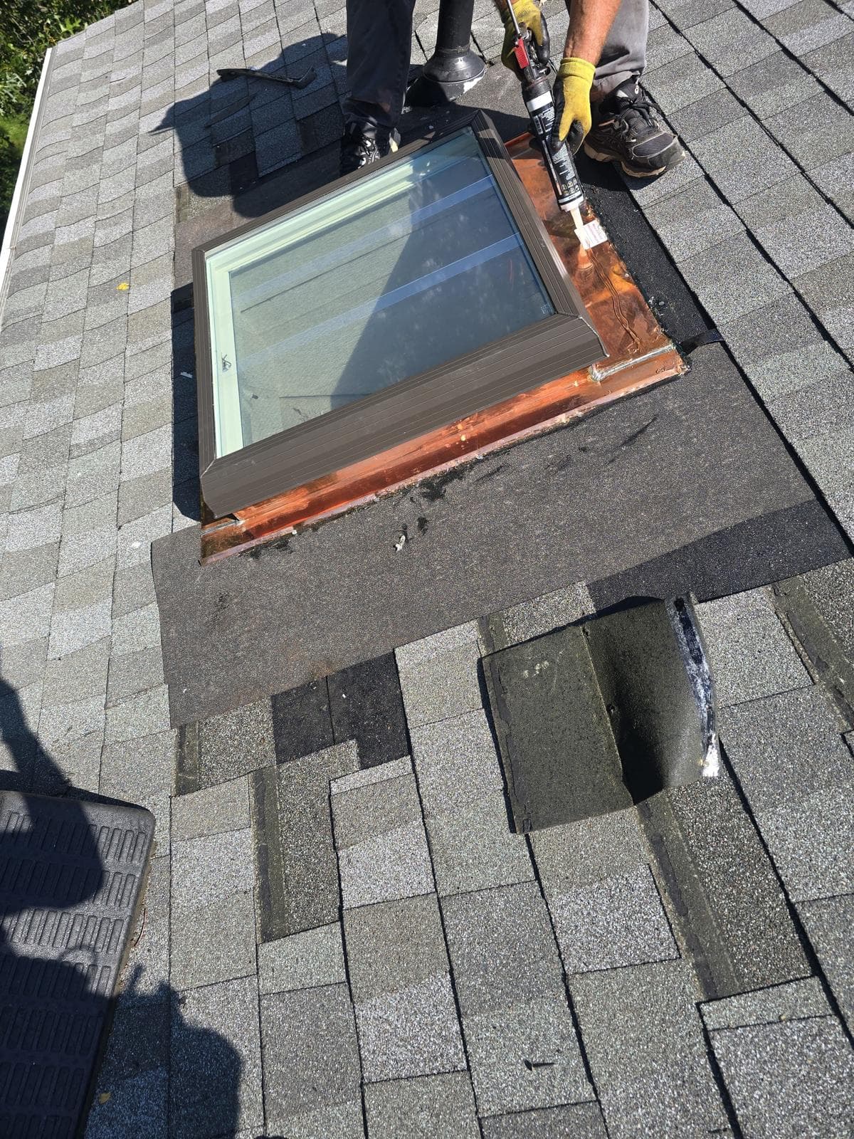 Roofer sealing copper flashing around a new skylight on a gray shingle roof